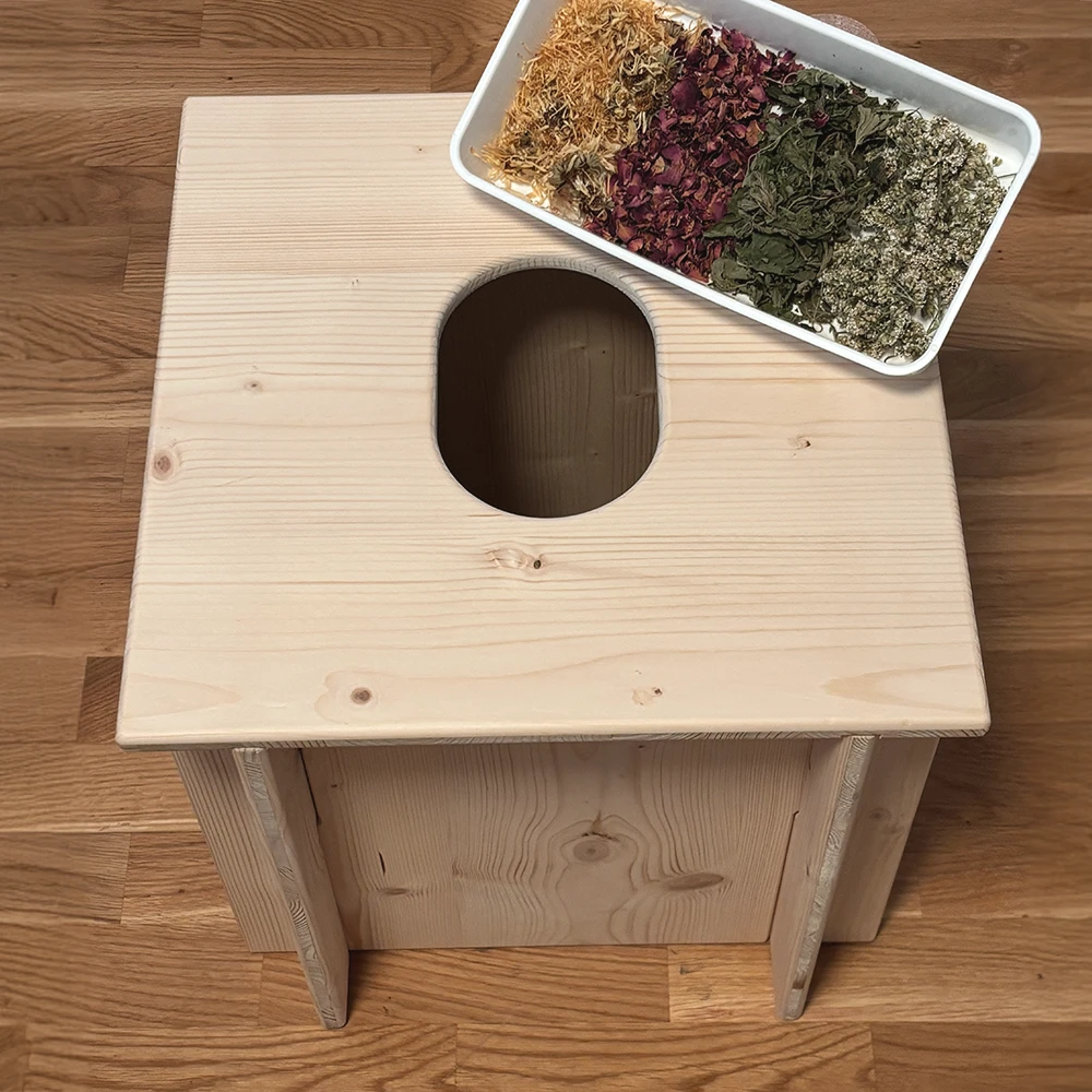 Holzstuhl für Yoni Steaming mit ovaler Öffnung, darauf eine Schale mit getrockneten Kräutern für die traditionelle Dampfanwendung