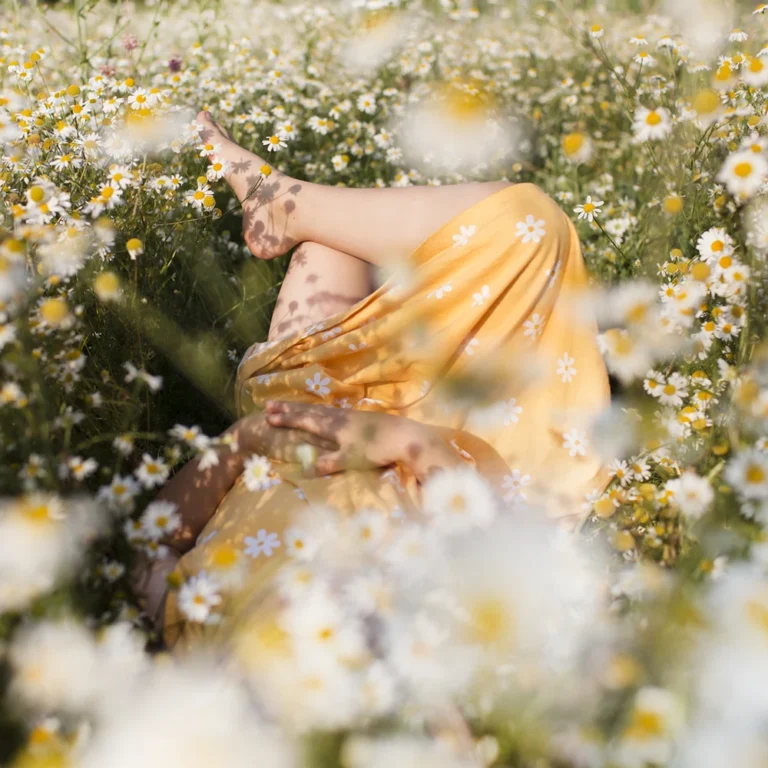Person in einem gelben Kleid liegt entspannt in einer blühenden Kamillenwiese, umgeben von weißen Blüten im warmen Sonnenlicht